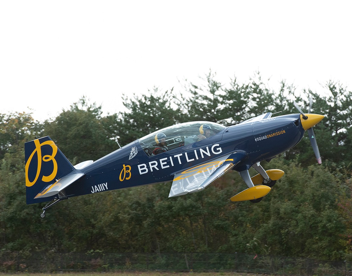 {generated} Breitlingのロゴを配した曲技飛行機が青黄の機体色でパイロットと共に森林背景の上を飛び立つ様子